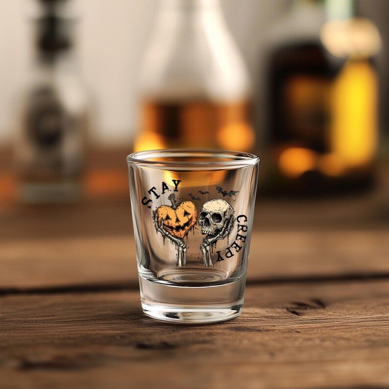 May include: A clear shot glass with a Halloween-themed design. The design features a jack-o'-lantern heart and a skull with the words "STAY CREEPY" printed on the glass. The glass is set on a wooden surface.