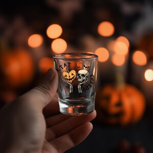 May include: A clear shot glass with a Halloween-themed design. The glass features a heart-shaped pumpkin and a skull, with the words "STAY CREEPY" printed on it. The background is blurred with orange bokeh lights.
