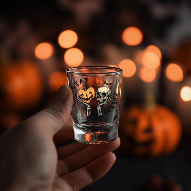May include: A clear shot glass with a Halloween-themed design. The glass features a heart-shaped pumpkin and a skull, with the words "STAY CREEPY" printed on it. The background is blurred with orange bokeh lights.