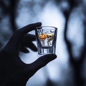 May include: Clear shot glass with a Halloween-themed design. The glass features a jack-o'-lantern and a skull, with the words "Stay Creepy" printed on the glass. Bats are also included in the design.