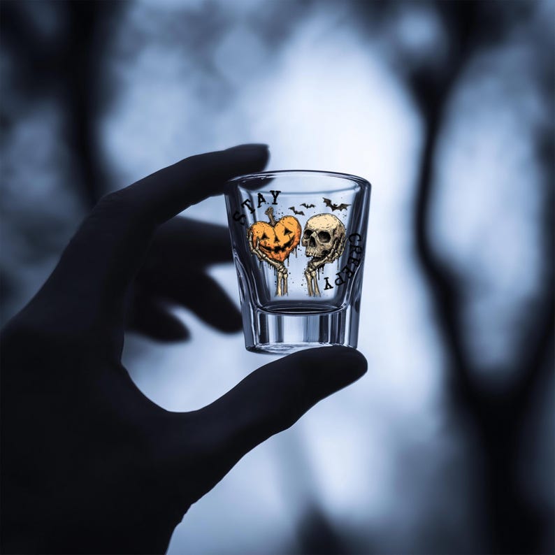 May include: Clear shot glass with a Halloween-themed design. The glass features a jack-o'-lantern and a skull, with the words "Stay Creepy" printed on the glass. Bats are also included in the design.