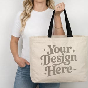 May include: Beige tote bag with black handles. The bag features the text "Your Design Here" in a stylized font. The bag is held by a person wearing a white shirt and blue jeans.