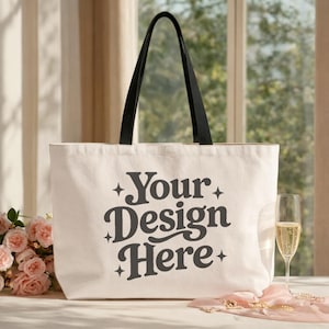 May include: A large, off-white tote bag with black handles. The bag features the text "Your Design Here" in a stylized, dark gray font. The bag is on a white surface, with a glass of champagne and pink roses in the background.
