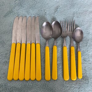 Puede incluir: Un juego de cubiertos de acero inoxidable con mangos de color amarillo brillante. El juego incluye cuchillos, cucharas y tenedores. Los cuchillos tienen hojas dentadas. Los cubiertos están colocados sobre una superficie texturizada azul claro.