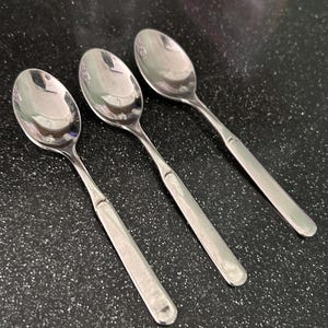May include: Three stainless steel spoons on a black speckled surface. The spoons have oval bowls and elongated, slightly flattened handles. The spoons reflect the light, creating a shiny appearance. The spoons are made of stainless steel.