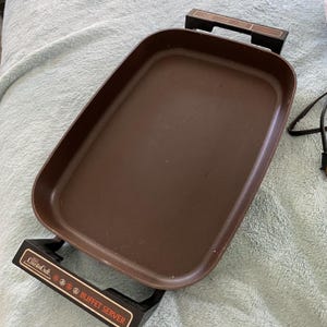 May include: Brown electric buffet server with black handles and the text "Counter Craft Buffet Server". The rectangular serving tray is brown and has a non-stick surface. The handles have a wood-grain design.