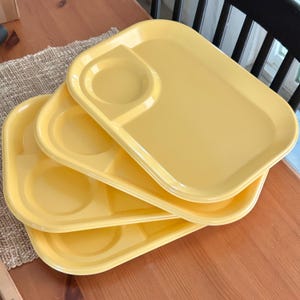 May include: A stack of four yellow plastic trays. Each tray has a rectangular shape with rounded corners, a circular indentation, and two divided sections. The trays are stacked on a wooden table.