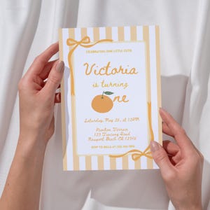 May include: A birthday invitation with a light orange and white striped background. The invitation features the text "Victoria is turning one" with an illustration of an orange. The invitation is held by two hands.