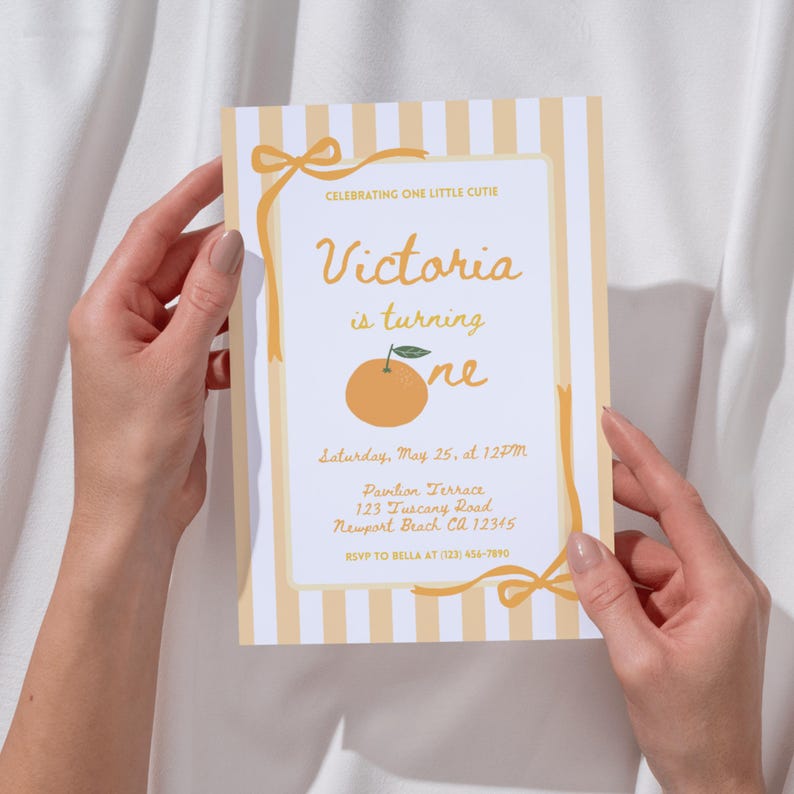 May include: A birthday invitation with a light orange and white striped background. The invitation features the text "Victoria is turning one" with an illustration of an orange. The invitation is held by two hands.