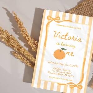 May include: A birthday invitation with a light orange and white striped background. The invitation features the text "Victoria is turning one" with an orange illustration. The invitation also includes event details and RSVP information.