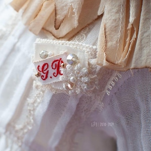 Pu&ograve; includere: Tessuto di pizzo bianco con una piccola etichetta ricamata in rosso che dice "G.K." circondata da perline bianche.