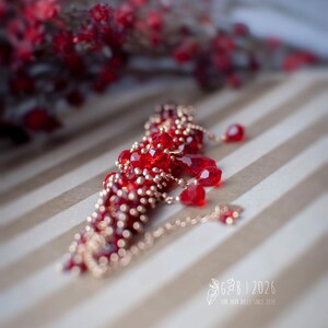 Może przedstawiać: Czerwony naszyjnik z koralik&oacute;w ze złotymi akcentami i delikatnym łańcuszkiem. Naszyjnik posiada fasetowane czerwone koraliki i mniejsze złote koraliki. Tło to wz&oacute;r w paski z rozmytym elementem kwiatowym.