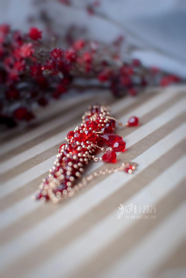 Może przedstawiać: Czerwony naszyjnik z koralik&oacute;w ze złotymi akcentami i delikatnym łańcuszkiem. Naszyjnik posiada fasetowane czerwone koraliki i mniejsze złote koraliki. Tło to wz&oacute;r w paski z rozmytym elementem kwiatowym.