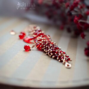 Może przedstawiać: Czerwona bransoletka z koralik&oacute;w ze złotymi akcentami i zawieszkami w kształcie łezki. Bransoletka jest prezentowana na paskowanej powierzchni, z rozmytymi czerwonymi jagodami w tle. Widoczny jest tekst "G&B | 2026 FOR YOUR DOLLS SINCE 2010".