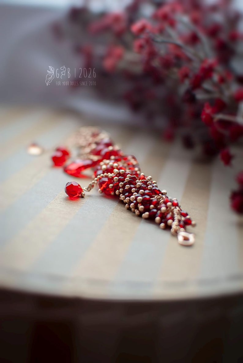 Może przedstawiać: Czerwona bransoletka z koralik&oacute;w ze złotymi akcentami i zawieszkami w kształcie łezki. Bransoletka jest prezentowana na paskowanej powierzchni, z rozmytymi czerwonymi jagodami w tle. Widoczny jest tekst "G&B | 2026 FOR YOUR DOLLS SINCE 2010".