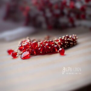 Może przedstawiać: Ręcznie robiony czerwony dodatek z koralik&oacute;w ze złotymi akcentami. Element posiada fasetowane czerwone koraliki i mniejsze złote koraliki, z wiszącymi czerwonymi kryształowymi kroplami. Widoczny jest tekst "G&B | 2026".