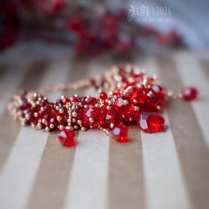 Może przedstawiać: Ręcznie robiona bransoletka z żywymi, czerwonymi, fasetowanymi koralikami i mniejszymi, złotymi koralikami. Bransoletka ma delikatny design z wiszącymi czerwonymi koralikami. Obraz zawiera tekst "G B | 2026 FOR YOUR DOLLS SINCE 2010".