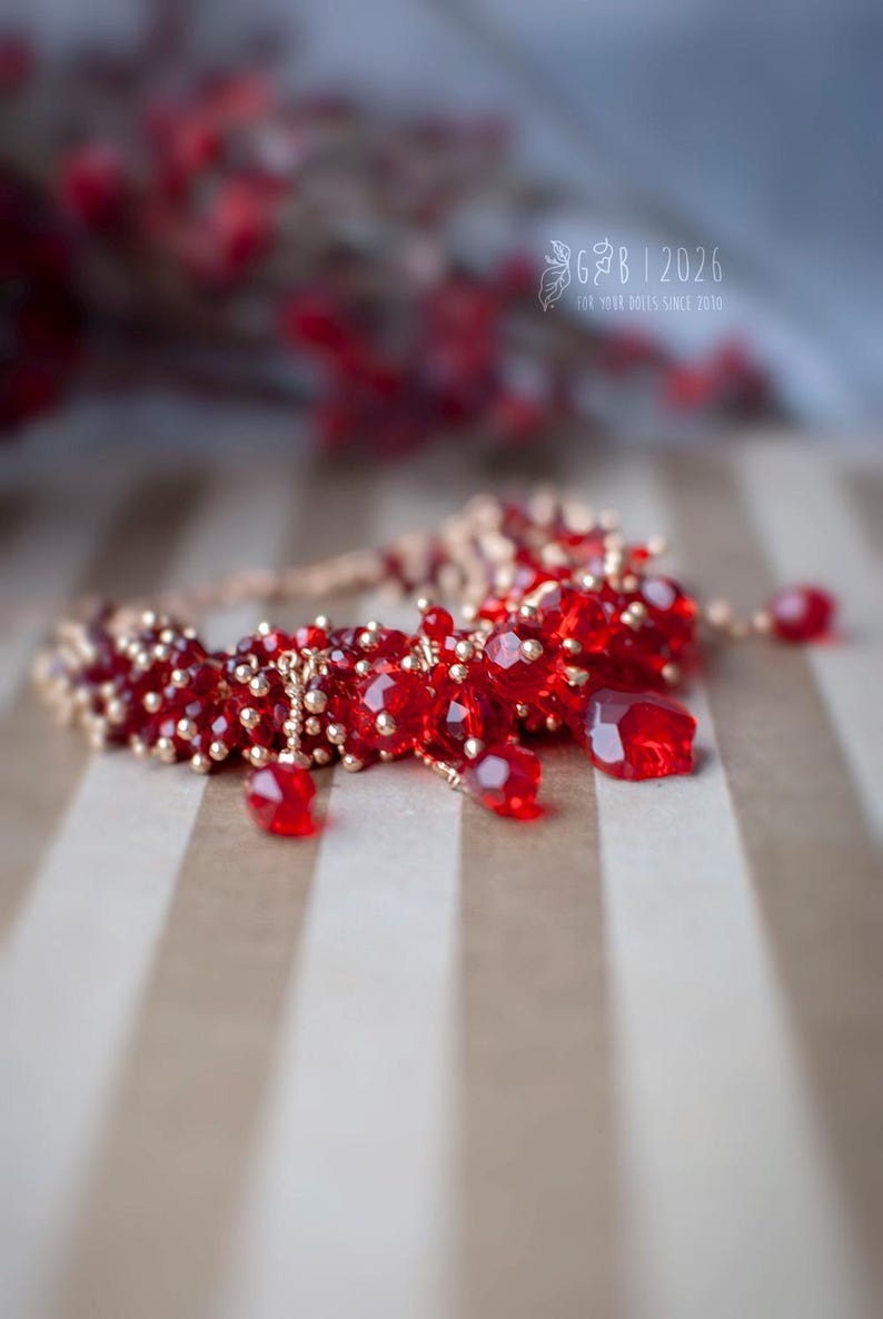 Może przedstawiać: Ręcznie robiona bransoletka z żywymi, czerwonymi, fasetowanymi koralikami i mniejszymi, złotymi koralikami. Bransoletka ma delikatny design z wiszącymi czerwonymi koralikami. Obraz zawiera tekst "G B | 2026 FOR YOUR DOLLS SINCE 2010".