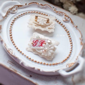 Pu&ograve; includere: Due spille in pizzo bianco con dettagli dorati. Una spilla presenta un motivo floreale con strass e una spilla di sicurezza. L'altra spilla ha le iniziali "G.H." ricamate in filo rosso.