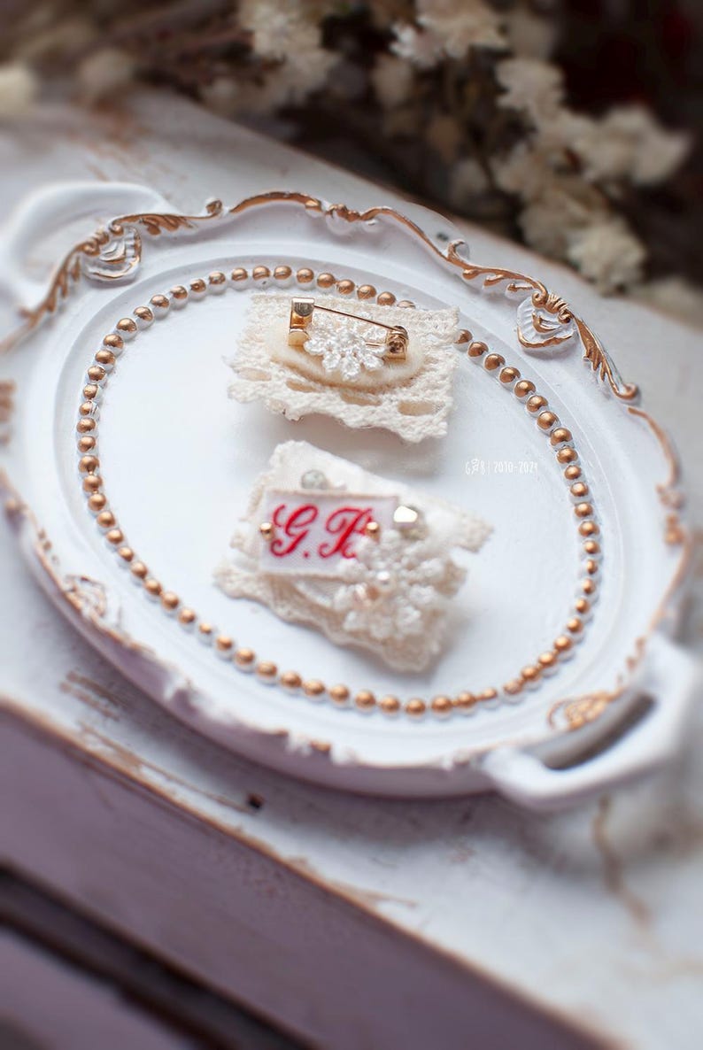 Pu&ograve; includere: Due spille in pizzo bianco con dettagli dorati. Una spilla presenta un motivo floreale con strass e una spilla di sicurezza. L'altra spilla ha le iniziali "G.H." ricamate in filo rosso.