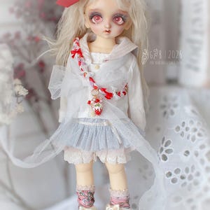 May include: A doll with long blonde hair and red eyes, wearing a white top, a blue skirt, and white shoes with bows. A red bow adorns the doll's head. The doll has a necklace with a strawberry pendant. The text "G&B | 2026" is visible.