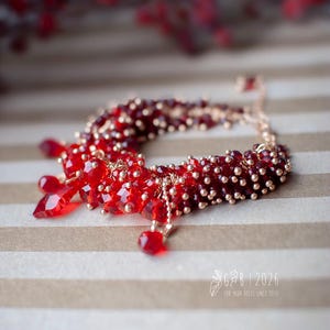 Może przedstawiać: Czerwona bransoletka z koralik&oacute;w ze złotymi akcentami. Bransoletka posiada skupisko czerwonych koralik&oacute;w i wiszące czerwone kryształowe zawieszki. Zawiera małe złote koraliki i delikatny łańcuszek. Widoczny jest tekst "GB | 2026".