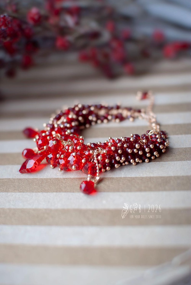 Może przedstawiać: Czerwona bransoletka z koralik&oacute;w ze złotymi akcentami. Bransoletka posiada skupisko czerwonych koralik&oacute;w i wiszące czerwone kryształowe zawieszki. Zawiera małe złote koraliki i delikatny łańcuszek. Widoczny jest tekst "GB | 2026".