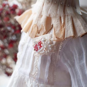 Pu&ograve; includere: Abito bianco in pizzo con colletto a balze e una piccola toppa ricamata che dice "G.F."