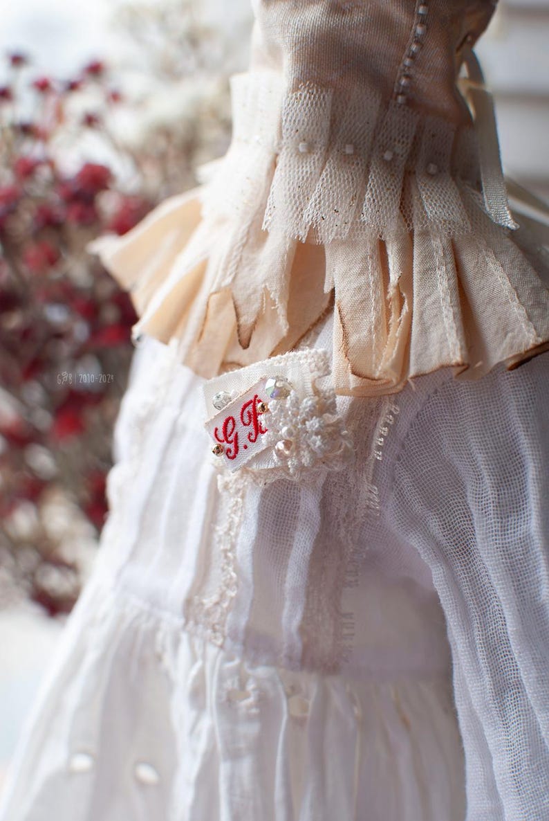 Pu&ograve; includere: Abito bianco in pizzo con colletto a balze e una piccola toppa ricamata che dice "G.F."