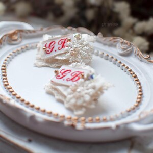 Pu&ograve; includere: Due decorazioni in pizzo e tessuto bianco con iniziali ricamate in rosso "G.F." e "G.B." su un vassoio ovale bianco con un bordo di perline dorate.