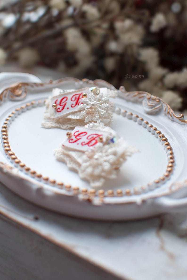 Pu&ograve; includere: Due decorazioni in pizzo e tessuto bianco con iniziali ricamate in rosso "G.F." e "G.B." su un vassoio ovale bianco con un bordo di perline dorate.