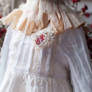 Pu&ograve; includere: Un vestito bianco con un bordo in pizzo e un colletto a balze. Il vestito ha una piccola toppa ricamata con le iniziali "G.F." in filo rosso.