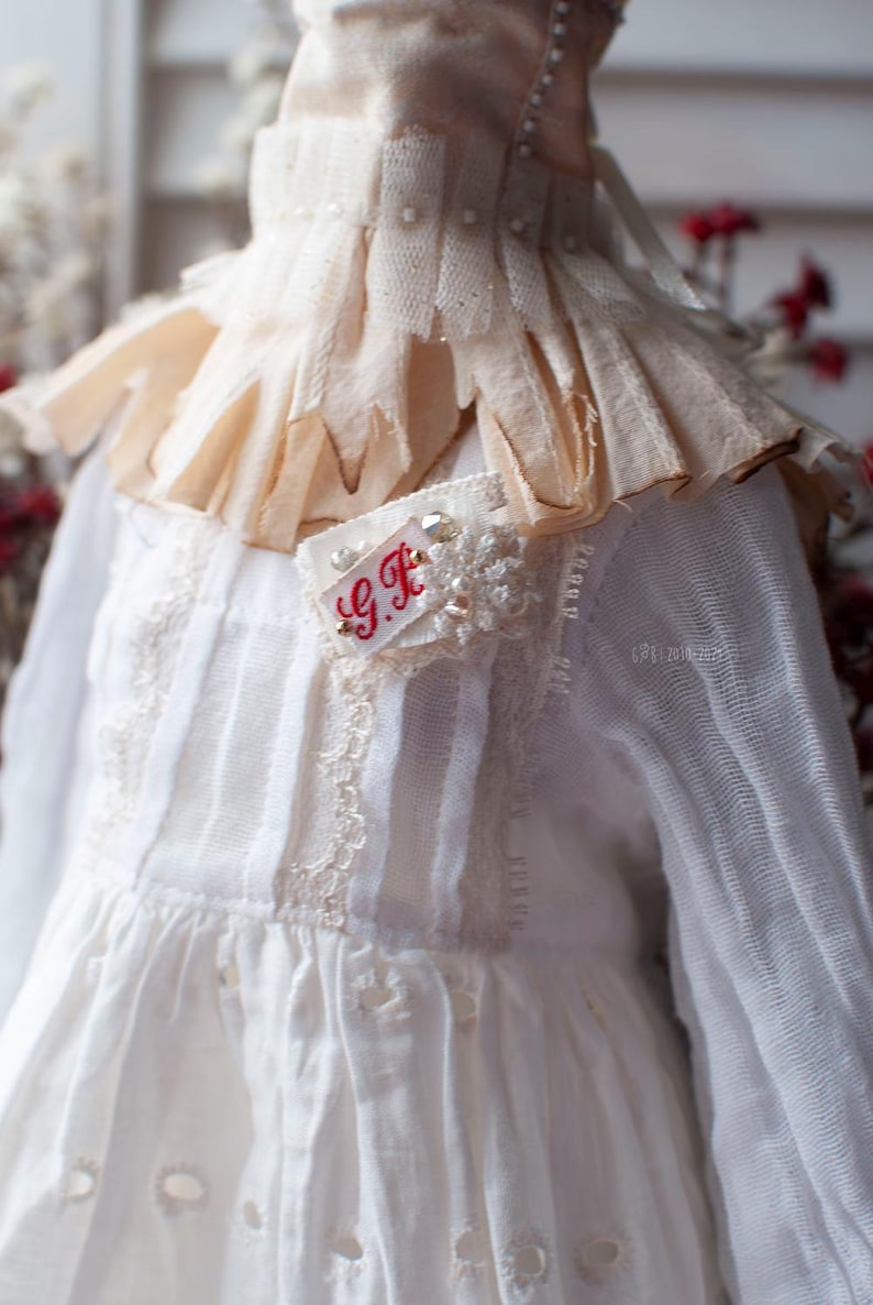 Pu&ograve; includere: Un vestito bianco con un bordo in pizzo e un colletto a balze. Il vestito ha una piccola toppa ricamata con le iniziali "G.F." in filo rosso.