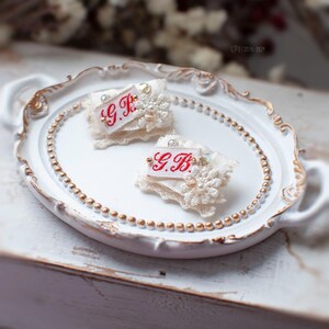 Pu&ograve; includere: Due spille in tessuto bianco con bordi in pizzo e dettagli di perle. Le spille sono su un vassoio bianco con un bordo di perline dorate. Le spille hanno ricamo rosso che dice "G.B."