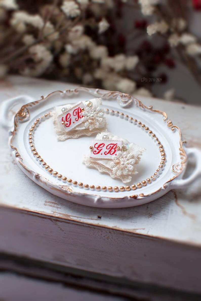 Pu&ograve; includere: Due spille in tessuto bianco con bordi in pizzo e dettagli di perle. Le spille sono su un vassoio bianco con un bordo di perline dorate. Le spille hanno ricamo rosso che dice "G.B."