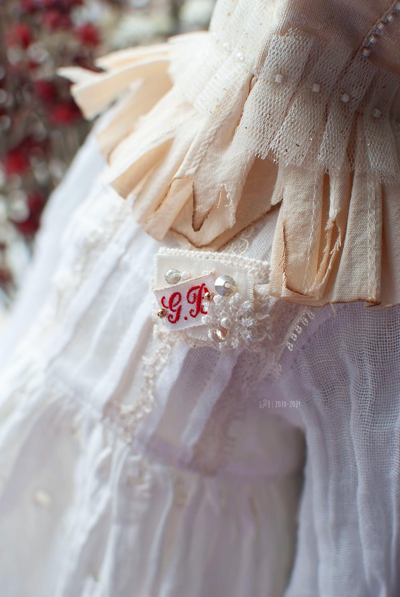 Pu&ograve; includere: Tessuto di pizzo bianco con una piccola etichetta ricamata in rosso che dice "G.K." circondata da perline bianche.