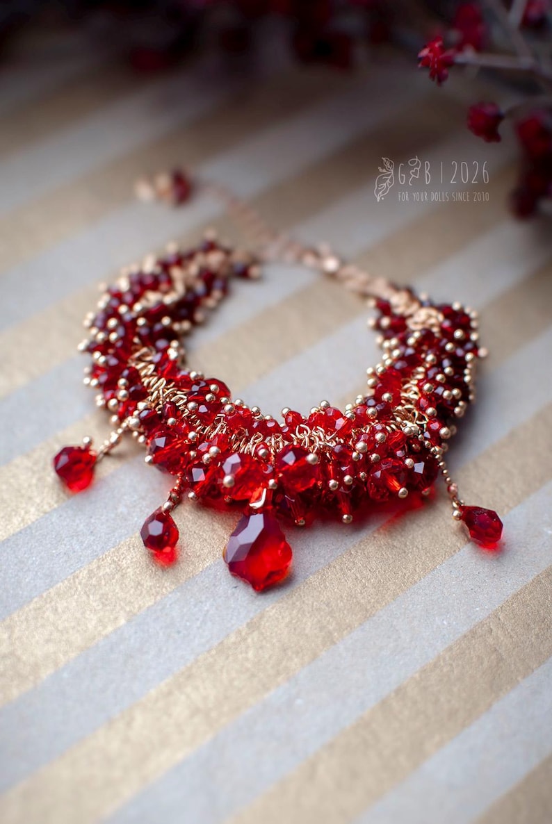 Może przedstawiać: Ręcznie robiona czerwona bransoletka ze złotymi akcentami. Bransoletka składa się z fasetowanych czerwonych koralik&oacute;w i mniejszych złotych koralik&oacute;w, z trzema czerwonymi zawieszkami w kształcie łezki. Tło to wz&oacute;r w złote i białe paski.