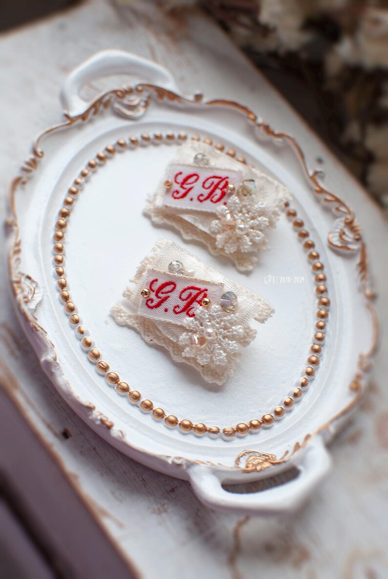 Pu&ograve; includere: Due pezzi di tessuto bianco con bordi in pizzo e dettagli di perle. Ogni pezzo ha un'etichetta ricamata in rosso con le iniziali "G.B."