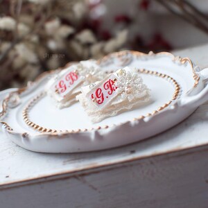 Pu&ograve; includere: Due decorazioni in pizzo bianco e perle con le iniziali ricamate in rosso "G.B." su un piatto bianco con bordo dorato.