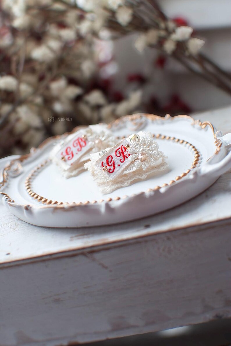 Pu&ograve; includere: Due decorazioni in pizzo bianco e perle con le iniziali ricamate in rosso "G.B." su un piatto bianco con bordo dorato.