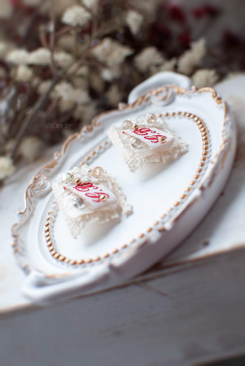 Pu&ograve; includere: Due piccoli quadrati di tessuto bianco con ricami rossi e bordatura in pizzo bianco. Ogni quadrato ha una piccola perla madreperla al centro ed &egrave; decorato con piccole perle bianche. I quadrati sono esposti su un vassoio bianco con un bordo di perline dorate.