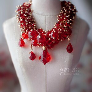 Może przedstawiać: Czerwony naszyjnik z koralik&oacute;w ze złotymi akcentami i wiszącymi czerwonymi kryształowymi zawieszkami. Naszyjnik jest prezentowany na białym manekinie. Odważny element biżuterii na specjalne okazje.