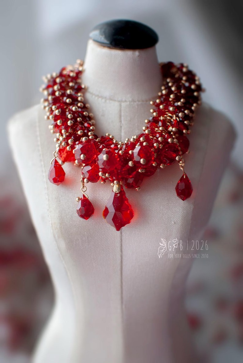 Może przedstawiać: Czerwony naszyjnik z koralik&oacute;w ze złotymi akcentami i wiszącymi czerwonymi kryształowymi zawieszkami. Naszyjnik jest prezentowany na białym manekinie. Odważny element biżuterii na specjalne okazje.