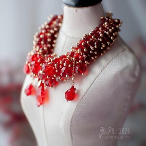 Może przedstawiać: Czerwony naszyjnik z koralik&oacute;w ze złotymi akcentami i kryształowymi zawieszkami, prezentowany na białym manekinie. Wyrazista biżuteria, idealna do dodania koloru.