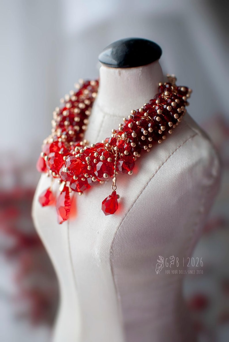 Może przedstawiać: Czerwony naszyjnik z koralik&oacute;w ze złotymi akcentami i kryształowymi zawieszkami, prezentowany na białym manekinie. Wyrazista biżuteria, idealna do dodania koloru.