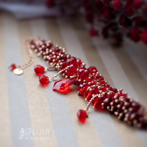 Może przedstawiać: Czerwony naszyjnik z koralik&oacute;w z wiszącymi czerwonymi kryształowymi zawieszkami. Naszyjnik posiada małe złote koraliki i delikatny łańcuszek. Tło obejmuje czerwone jagody i pasiastą powierzchnię. Widoczny jest tekst "G&B | 2026".