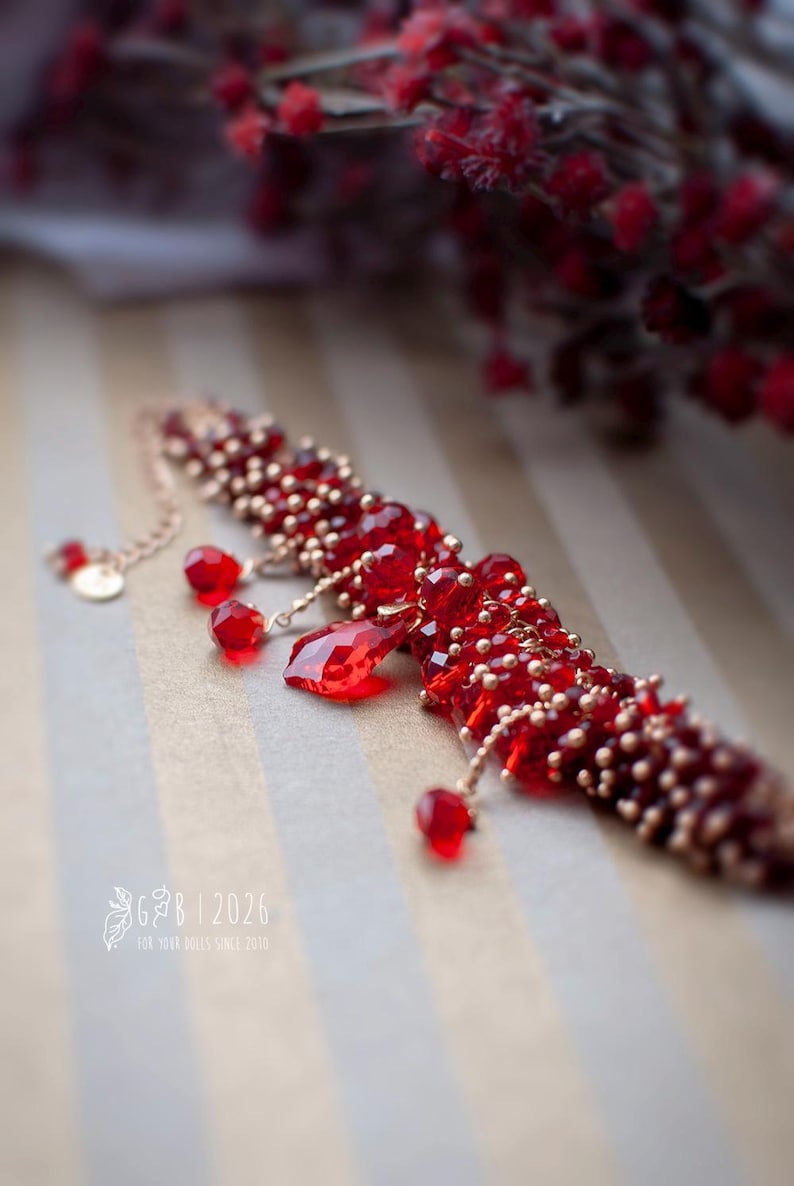 Może przedstawiać: Czerwony naszyjnik z koralik&oacute;w z wiszącymi czerwonymi kryształowymi zawieszkami. Naszyjnik posiada małe złote koraliki i delikatny łańcuszek. Tło obejmuje czerwone jagody i pasiastą powierzchnię. Widoczny jest tekst "G&B | 2026".