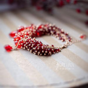 Może przedstawiać: Bransoletka z czerwonych i złotych koralik&oacute;w, składająca się z małych złotych i większych, głębokich czerwonych koralik&oacute;w. Bransoletka leży na paskowanej powierzchni. Obraz zawiera tekst "G&B | 2026" i "FOR YOUR SOLES SINCE 2010".