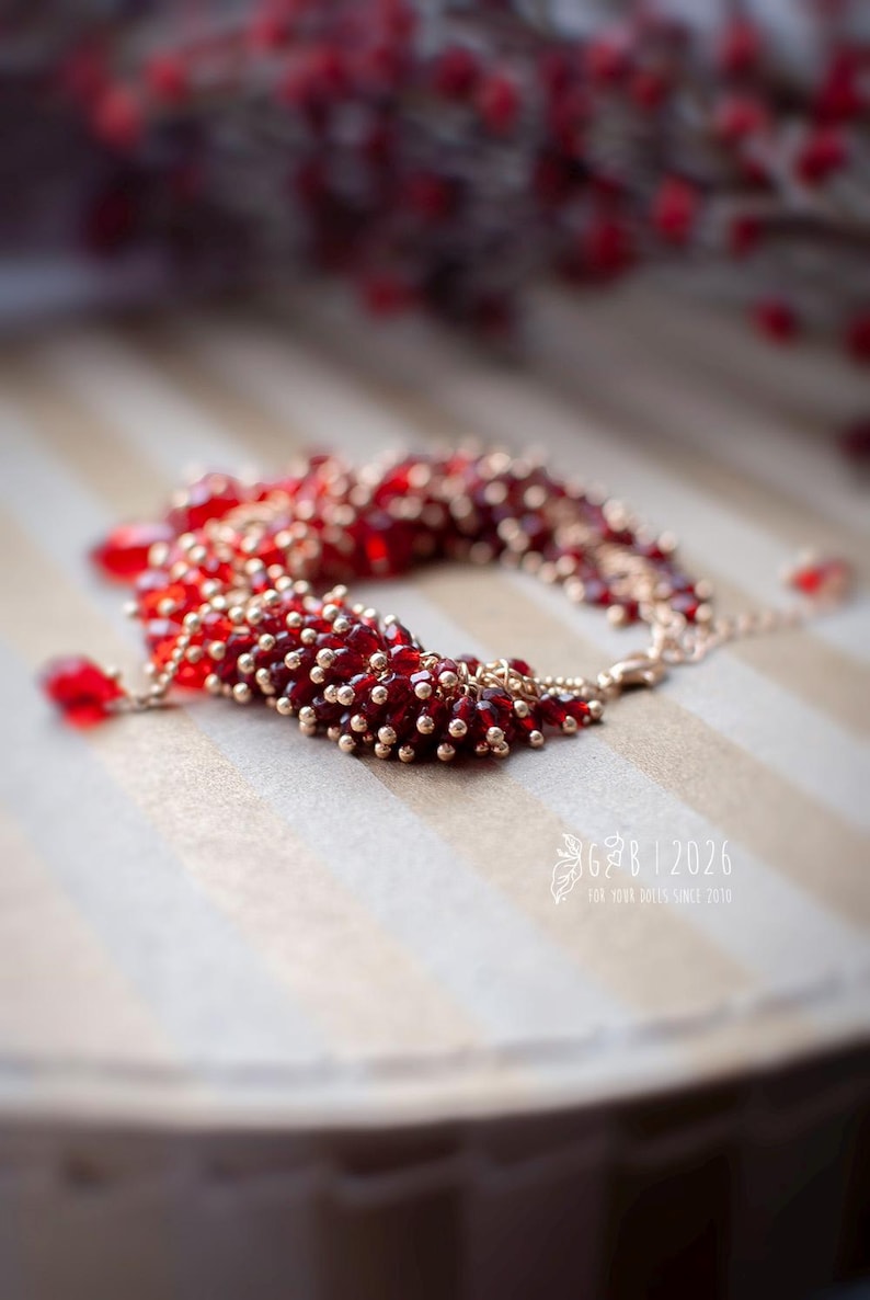 Może przedstawiać: Bransoletka z czerwonych i złotych koralik&oacute;w, składająca się z małych złotych i większych, głębokich czerwonych koralik&oacute;w. Bransoletka leży na paskowanej powierzchni. Obraz zawiera tekst "G&B | 2026" i "FOR YOUR SOLES SINCE 2010".