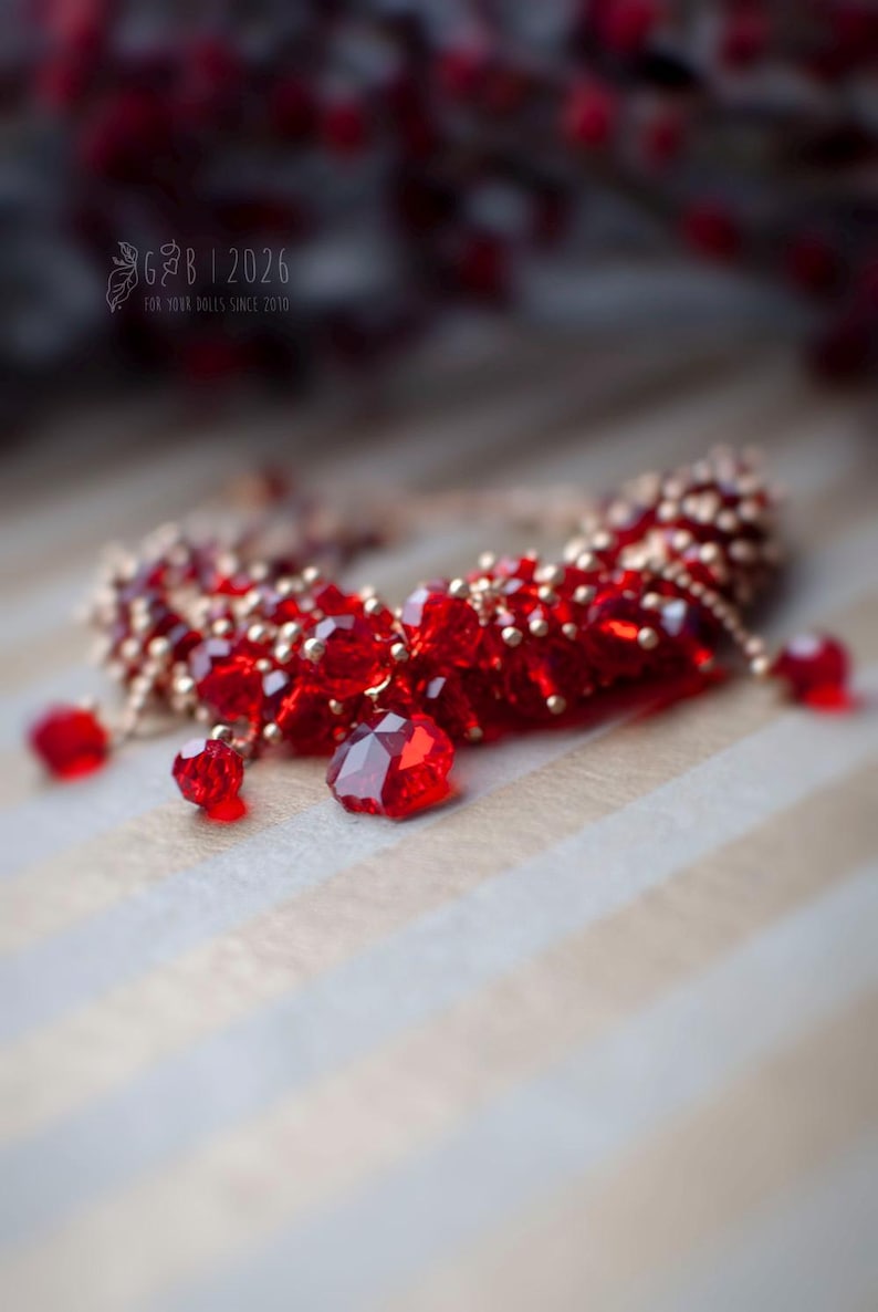 Może przedstawiać: Czerwony naszyjnik z koralik&oacute;w ze złotymi akcentami i wiszącymi czerwonymi kryształowymi zawieszkami. Biżuteria jest umieszczona na tle w paski, z rozmytym elementem kwiatowym w tle.